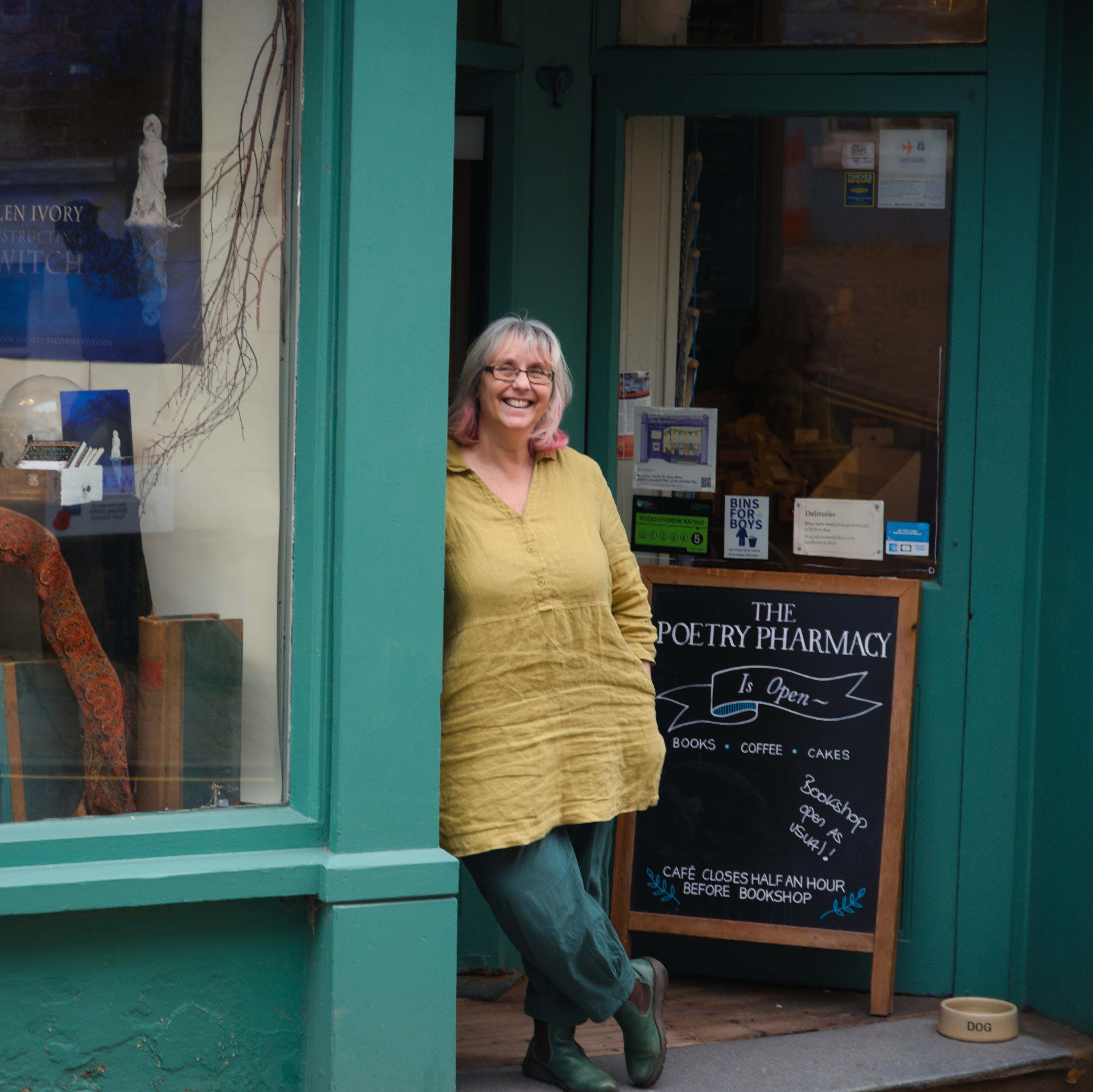 Deborah Alma standing outside a teal building with 'The Poetry Pharmacy' sign.